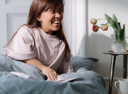 Asian woman with brown hair smiling, enjoying a calm morning in bed, surrounded by flowers and coffee.