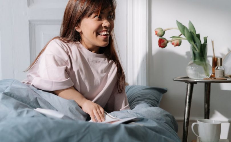 Asian woman with brown hair smiling, enjoying a calm morning in bed, surrounded by flowers and coffee.